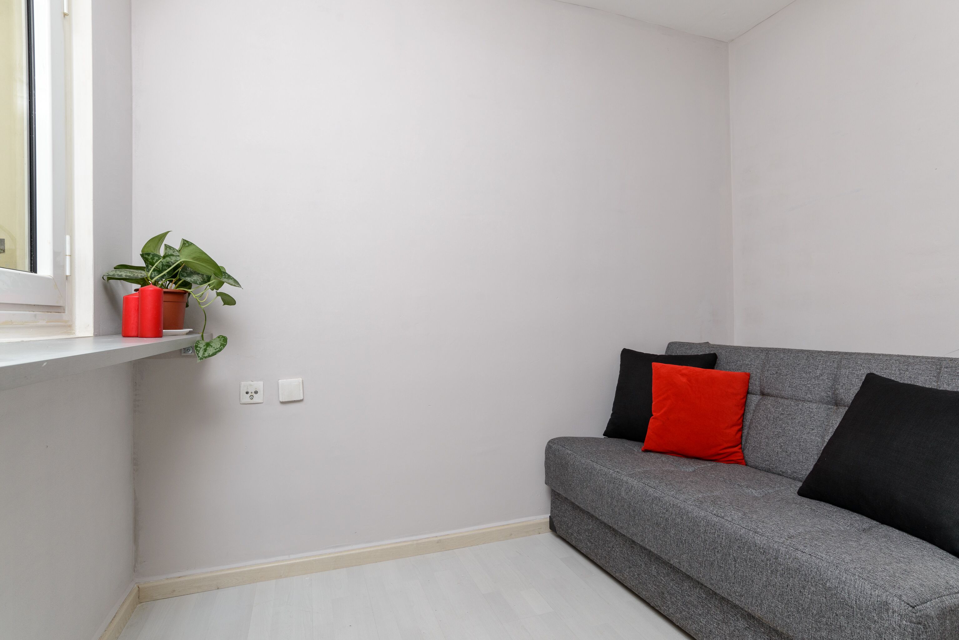 Quarto de canto com sofá cinza, almofada vermelha e planta em vaso sobre uma prateleira branca.