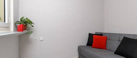 Quarto de canto com sofá cinza, almofada vermelha e planta em vaso sobre uma prateleira branca.