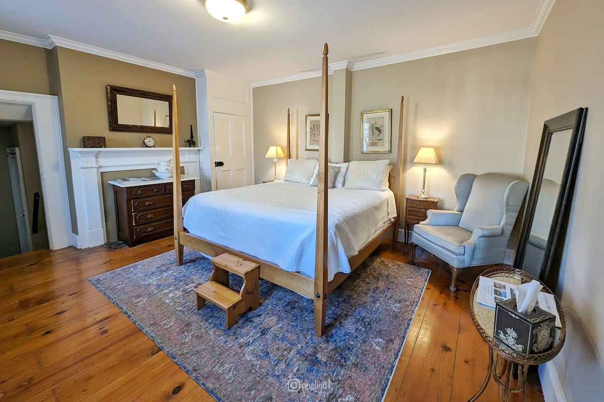 A four-poster bed with a wooden frame, a small wooden stool, a nightstand with a lamp, a chair, and a bedside table with a tissue box.