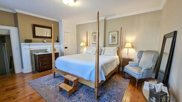 Una cama con dosel de madera, un taburete pequeño de madera, una mesita de noche con una lámpara, una silla y una mesilla con una caja de pañuelos.