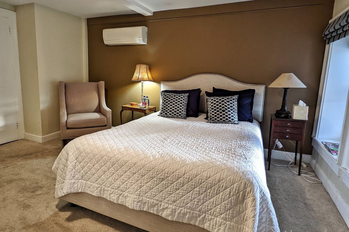 A hotel room with a large bed, a chair, a nightstand with a lamp, and an air conditioning unit on the wall.