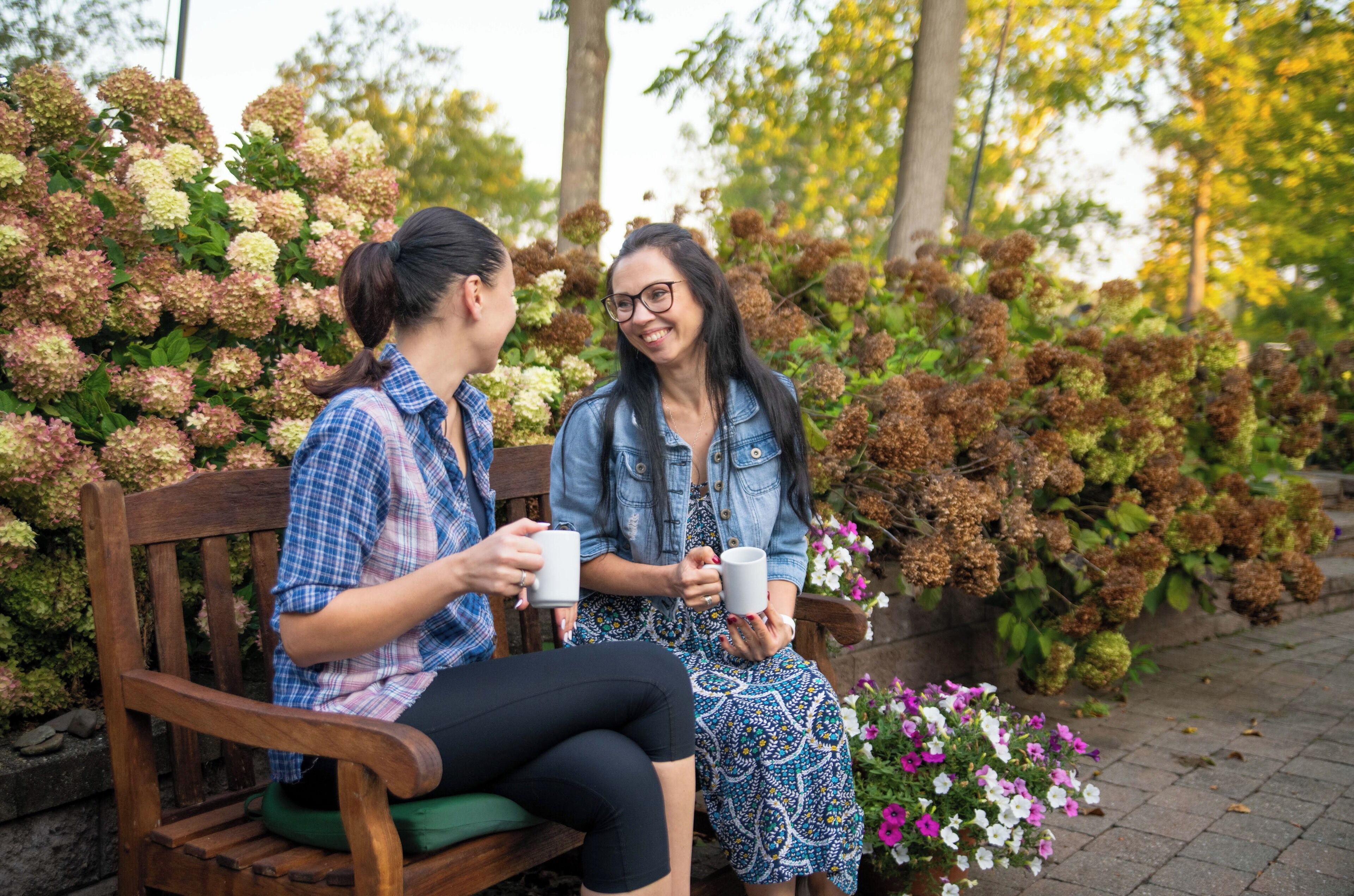 Two people sitting on a wooden bench outdoors, holding mugs.