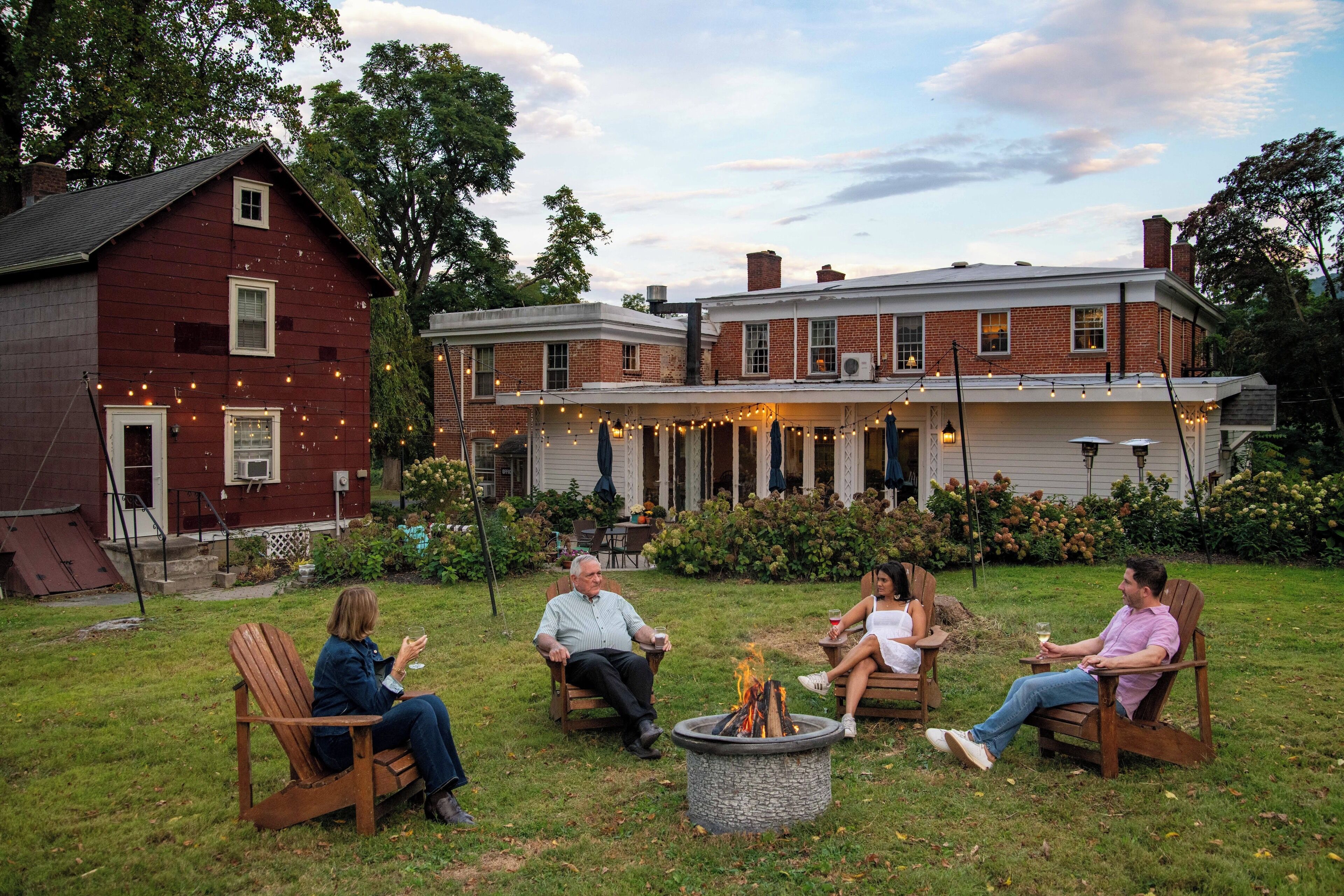 A group of people seated around a fire pit in a backyard with a brick house and a red building in the background.