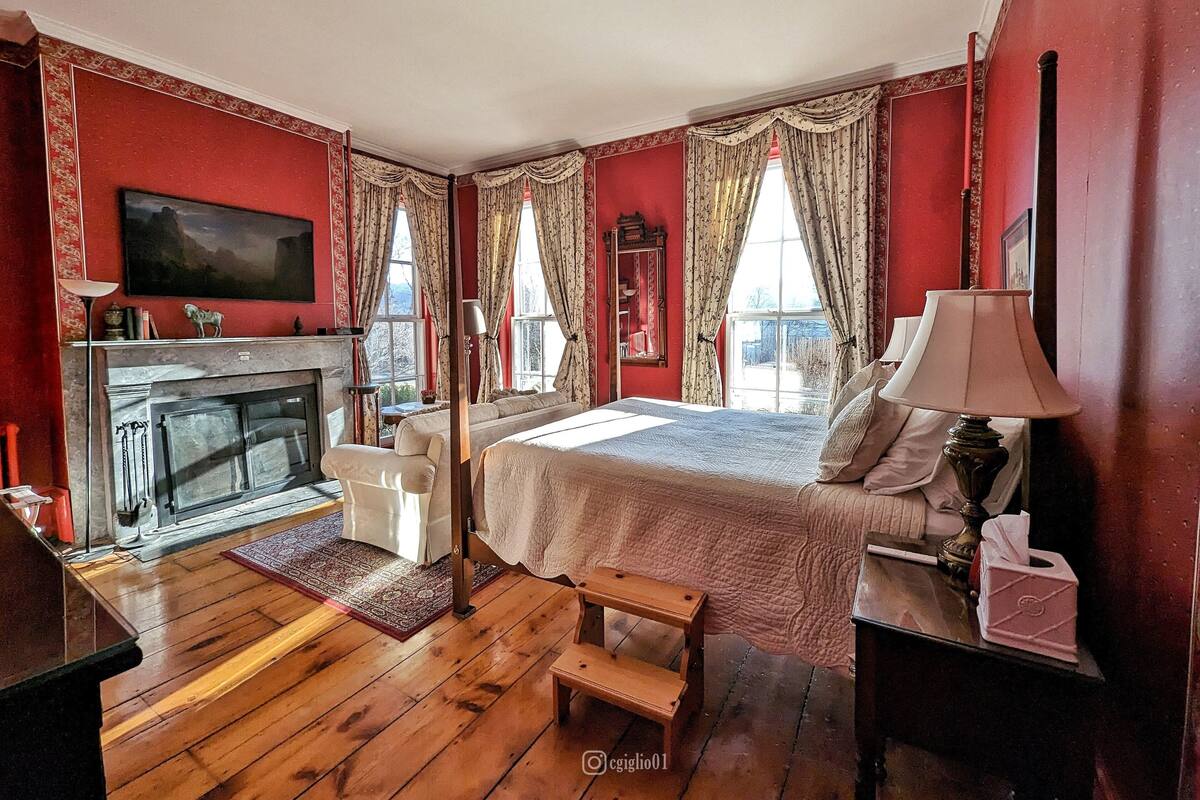 A bedroom with a fireplace, a bed, a desk, and a window with curtains.
