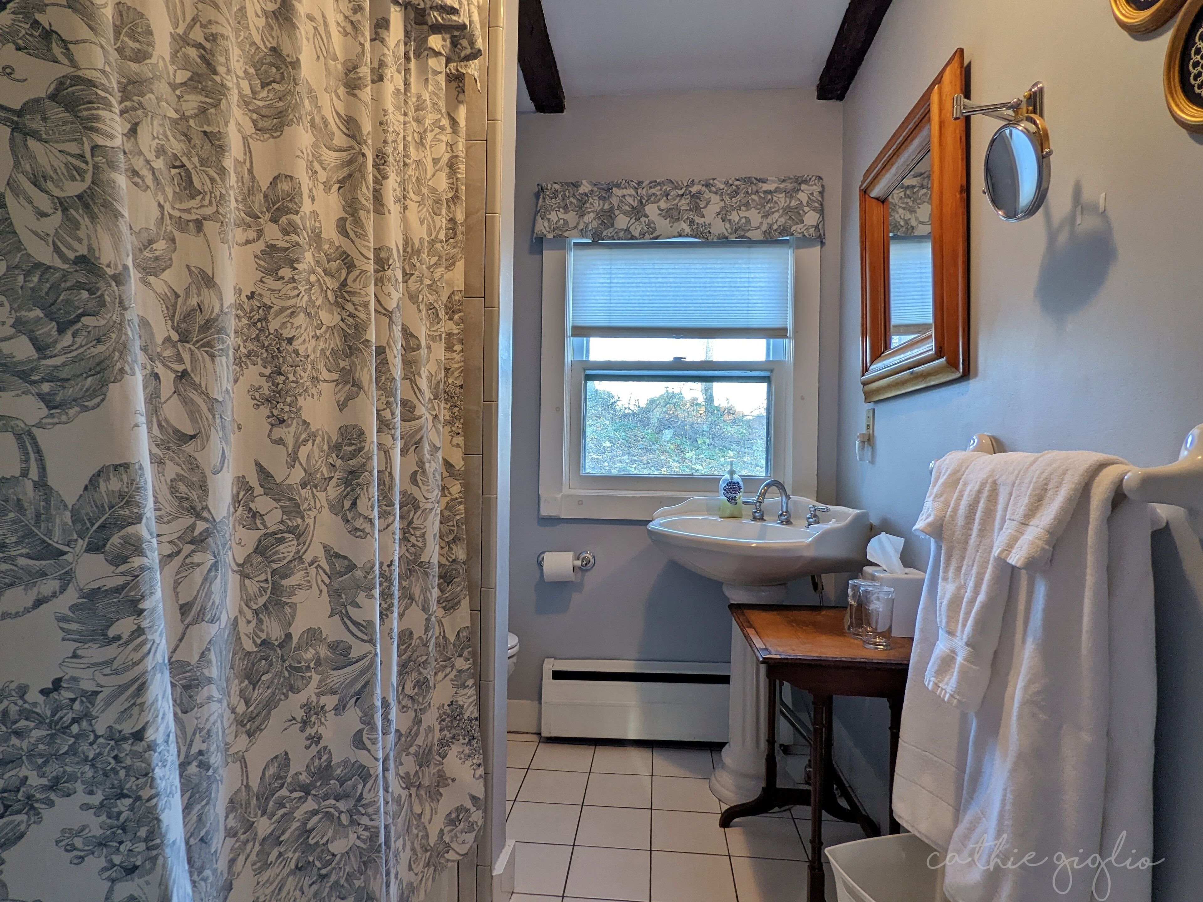 Une salle de bain avec un rideau à motifs floraux, une fenêtre avec des stores, un lavabo avec un miroir et une serviette accrochée au mur.