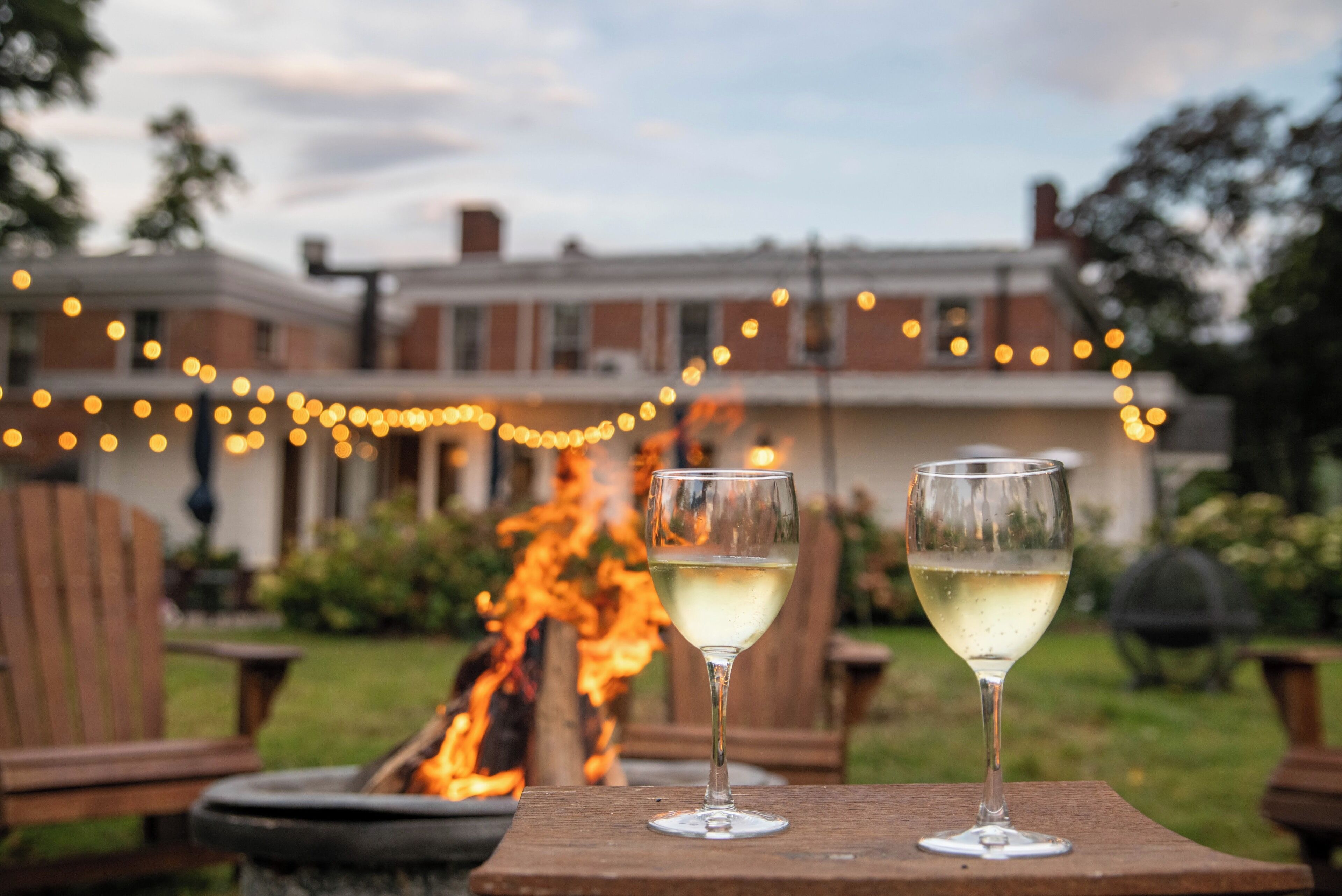 Two glasses of white wine on a wooden table with a fire in the background and a house with string lights.