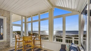 A covered outdoor seating area with a view of the ocean, wooden chairs, a sofa, and a striped rug.