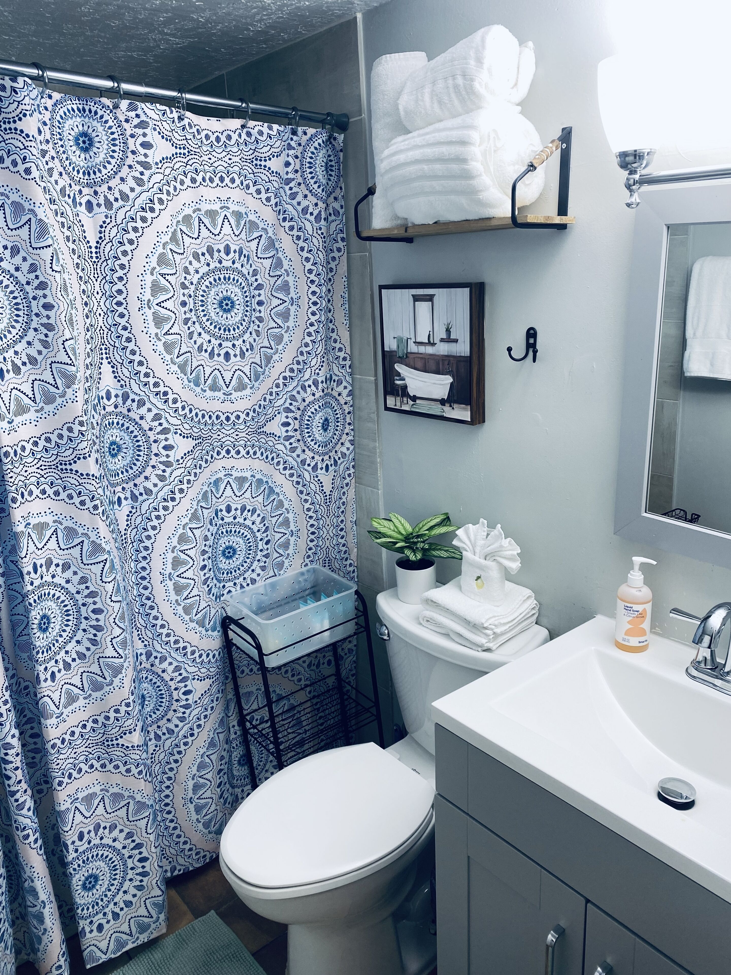 A bathroom with a patterned shower curtain, a toilet, a sink with a mirror, and a shelf with towels.