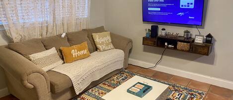 Ein Wohnzimmer mit braunem Sofa, gemusterten Kissen, weißer Decke, einem Couchtisch mit dekorativer Schachtel, einem farbenfrohen Teppich und einem an der Wand befestigten Flachbildfernseher.