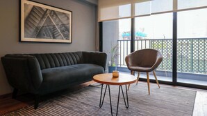 Ein modernes Wohnzimmer mit einer dunklen Couch, einem runden Holztisch und einem Sessel. Ein gerahmtes Bild hängt an der Wand und ein Blumentopf steht auf der Fensterbank.