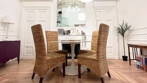 Een eetgedeelte met een witte tafel, twee rieten stoelen, een kroonluchter en een spiegel aan de muur.