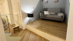 Een kamer met een grijze bank, een klein tafeltje, een lamp en een trap.