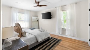 A bedroom with a bed, a ceiling fan, a TV mounted on the wall, and a window with curtains.