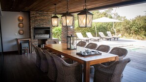 An outdoor dining area with a wooden table and wicker chairs, a stone wall, and a TV mounted on the wall. - Kruger National Elephant Point Lodge (Mbombela)