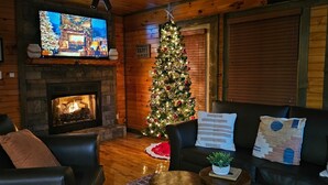 Un salon chaleureux avec une cheminée, un téléviseur, un sapin de Noël et un canapé agrémenté de coussins décoratifs.