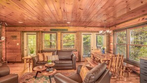 L’intérieur chaleureux d’un chalet avec un plafond en bois, un canapé en cuir, une table à manger et de grandes fenêtres équipées de stores.