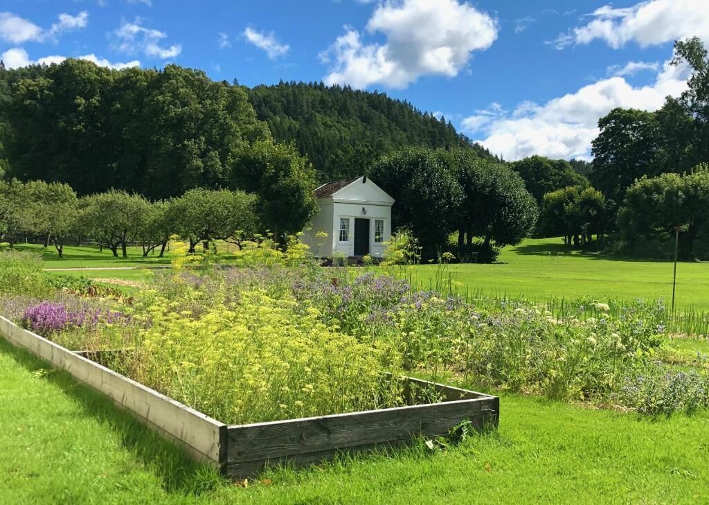 Ein kleines, weißes Gebäude mit einer grünen Tür, umgeben von einem gepflegten Garten mit verschiedenen Pflanzen und einem Holzzaun.