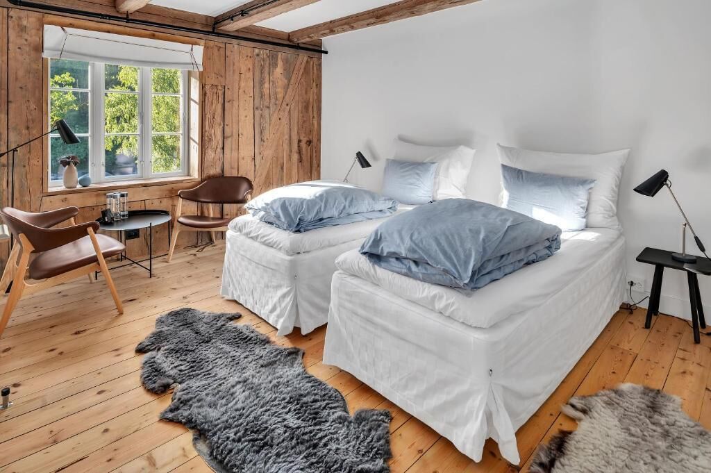 A room with two beds, a wooden floor, a large window, a chair, a table, and a fur rug.