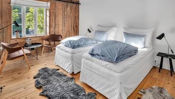 A room with two beds, a wooden floor, a large window, a chair, a table, and a fur rug.