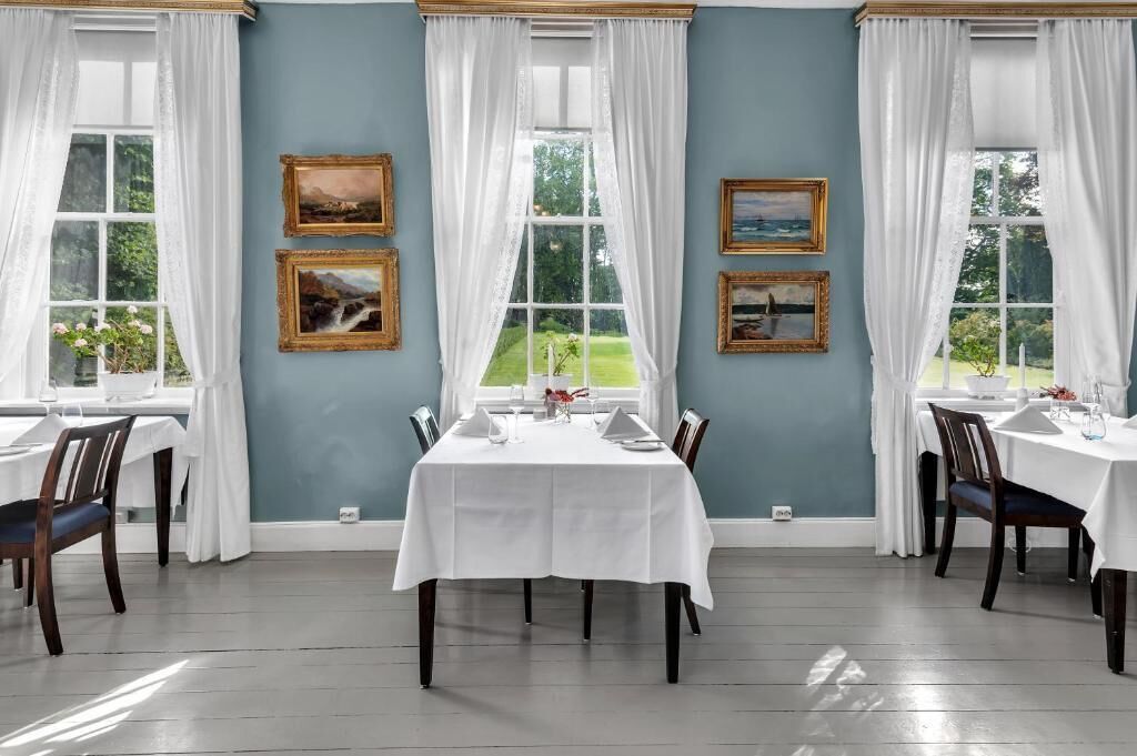A dining room with a white table set for four, large windows with white curtains, and framed landscape paintings on the walls.