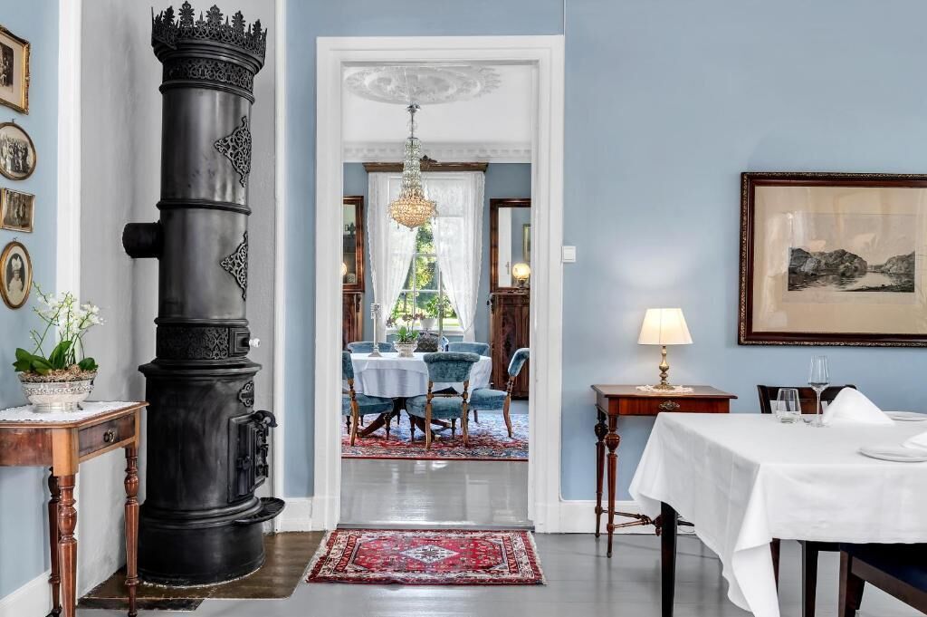 A hallway with a large black stove, a dining area with blue chairs, a chandelier, and a framed picture on the wall.