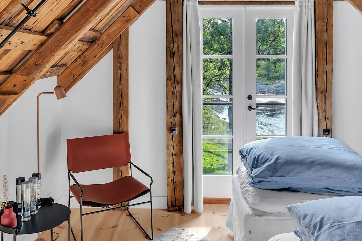 A cozy attic bedroom with a sloped wooden ceiling, a bed with white bedding, a red chair, a small table with candles, and a view of greenery through a glass door.