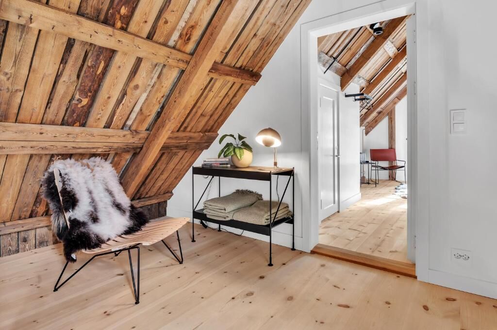 A loft-style room with exposed wooden beams, a bench, a small table with a lamp, and a door leading to another room.