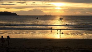 Una playa al atardecer con aguas tranquilas, barcos y gente caminando por la orilla.