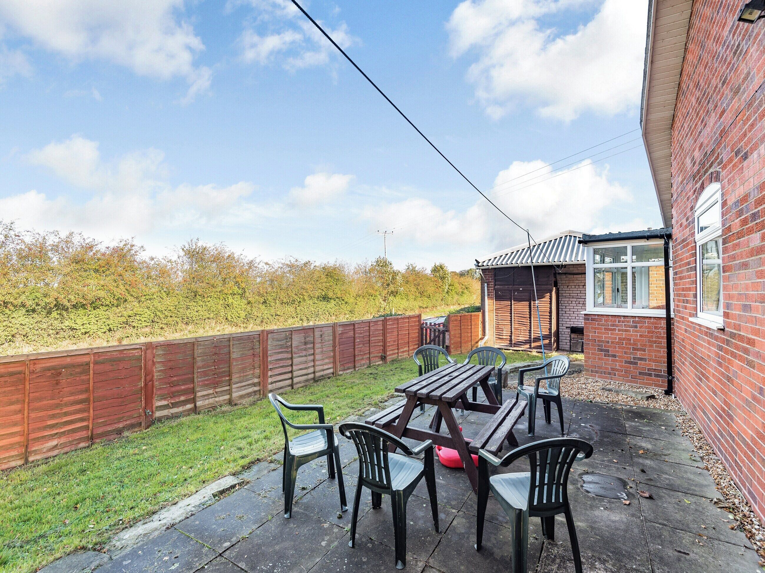 A patio area with a wooden table and chairs, a brick wall, and a view of a grassy field.