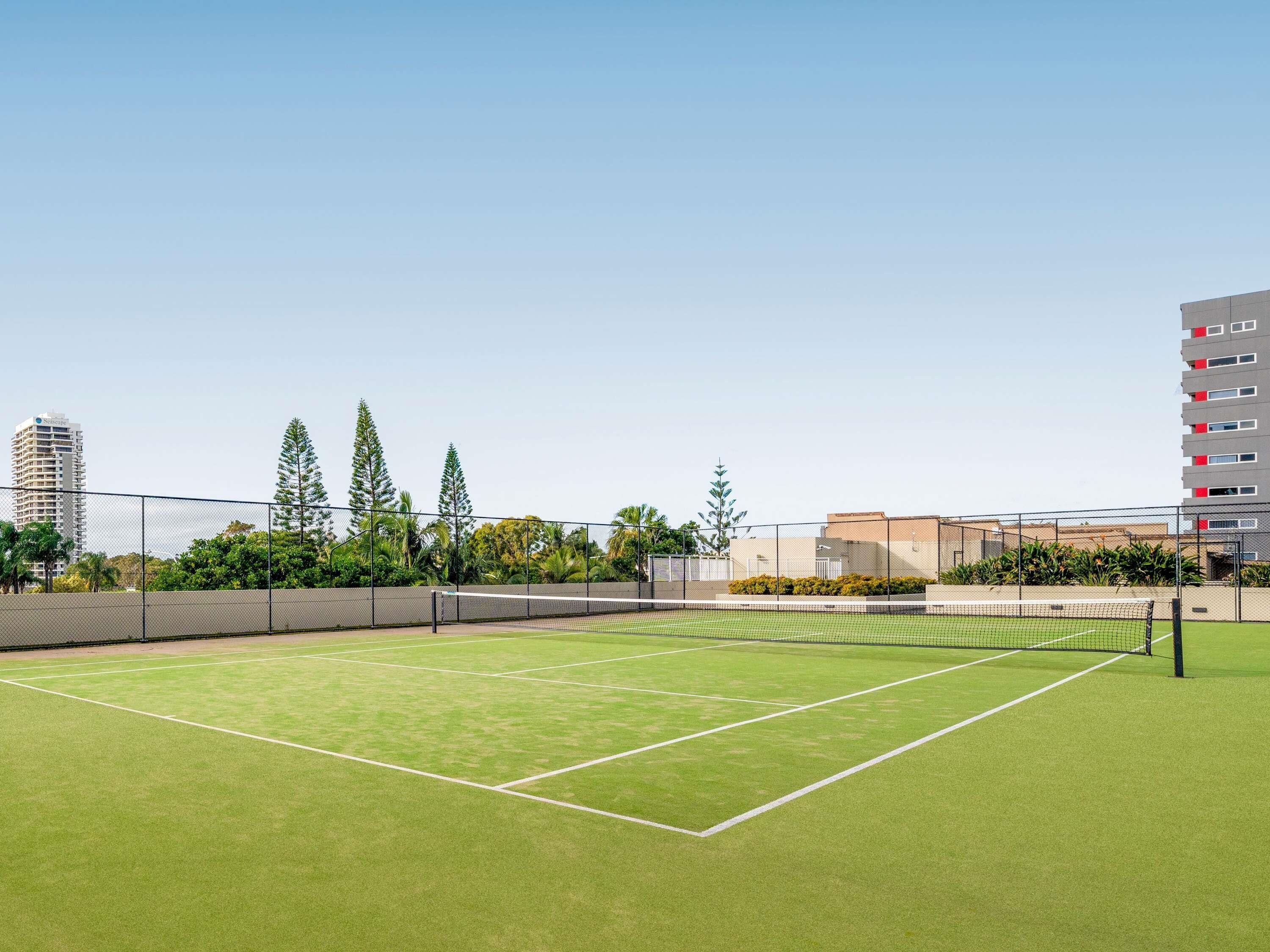 Una cancha de tenis al aire libre con una red, rodeada por una cerca, árboles y edificios al fondo.