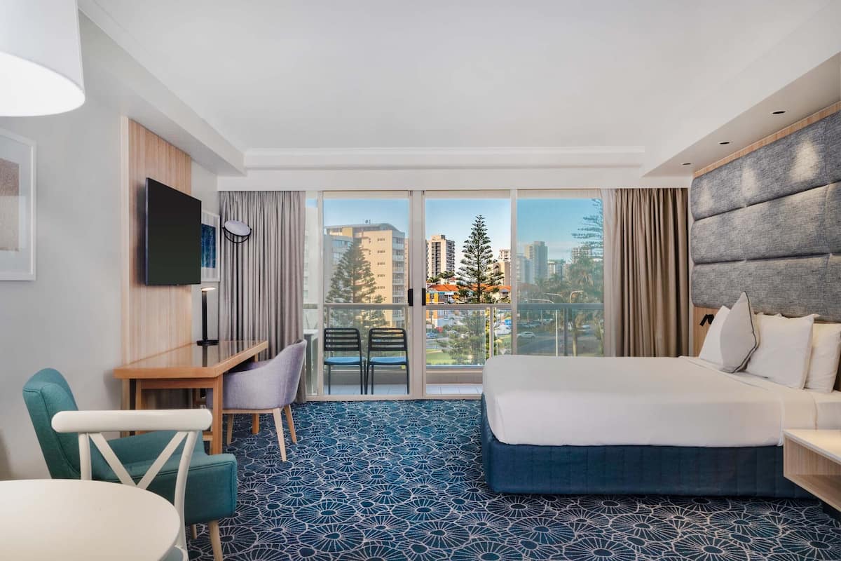 A hotel room with a bed, a desk, two chairs, a TV, and a large window with a city view.
