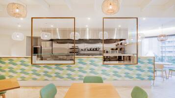 A modern restaurant with a geometric patterned wall, a central kitchen area, and green upholstered chairs.