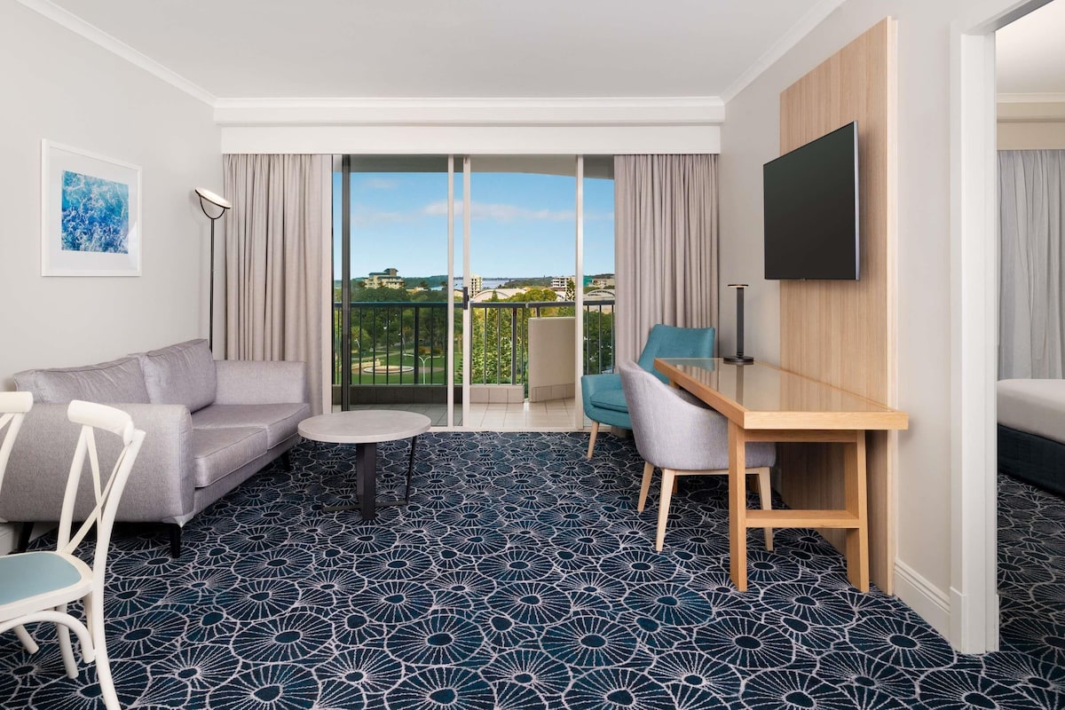 A hotel room with a sofa, a small round table, a desk with a chair, a TV mounted on the wall, and a balcony with a view of the city.