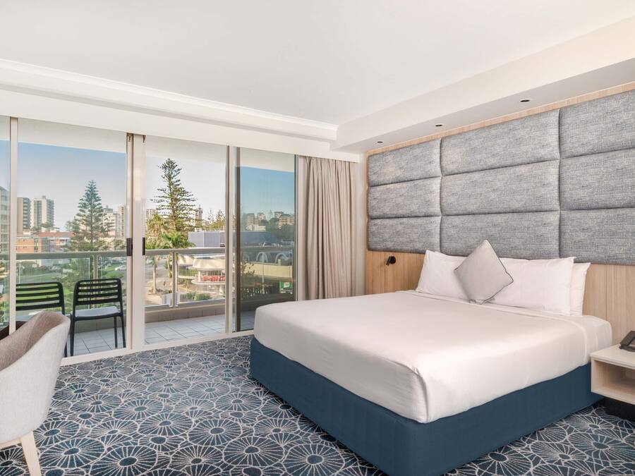 Une chambre d’hôtel avec un grand lit, une vue sur la ville, un balcon avec des chaises et un sol à motifs.