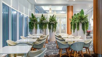A modern indoor dining area with wicker chairs and white tables, surrounded by vertical blinds and potted plants.