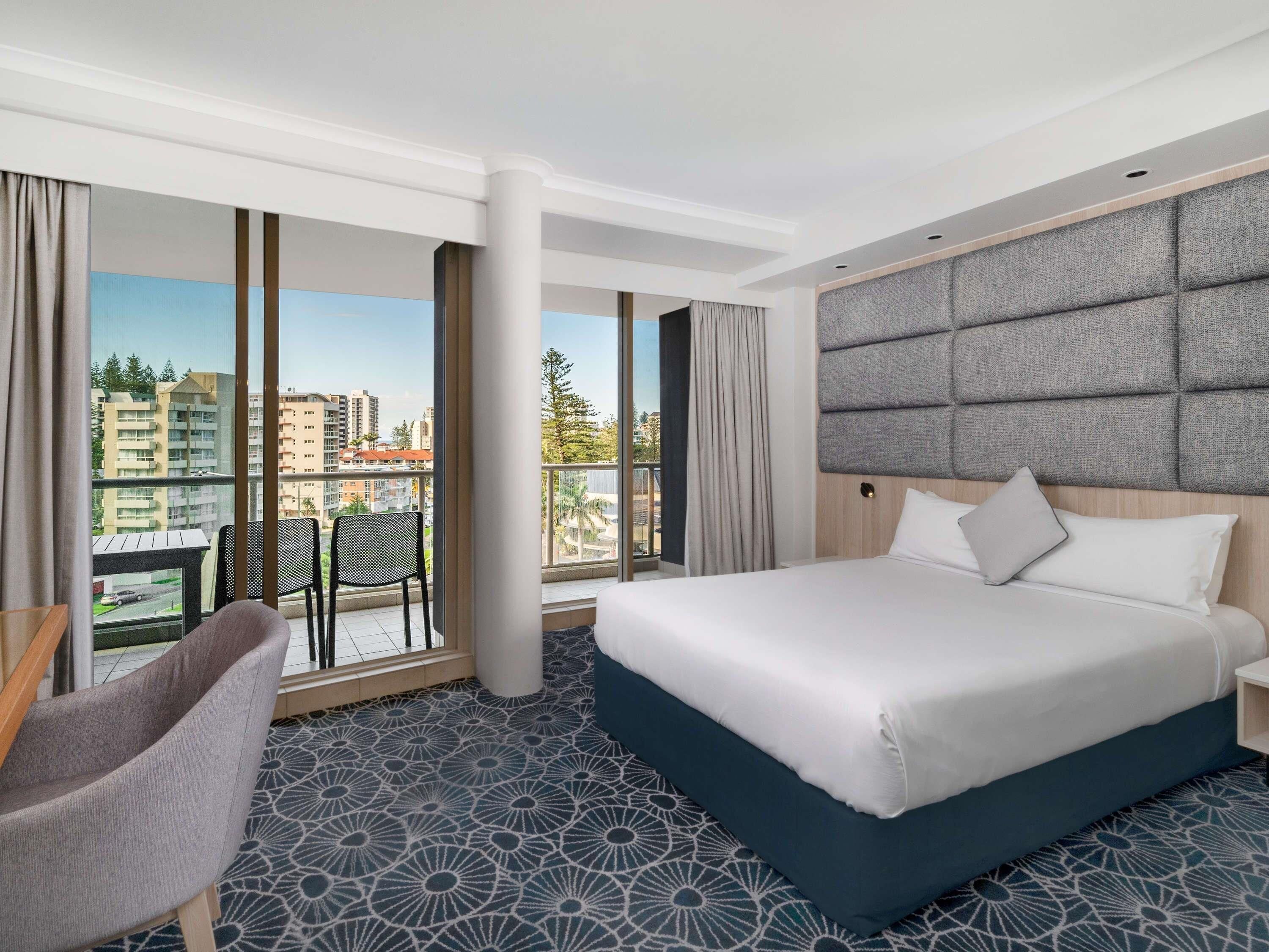 Quarto de hotel com uma cama grande, uma poltrona, uma escrivaninha e vista para a cidade através de grandes janelas.