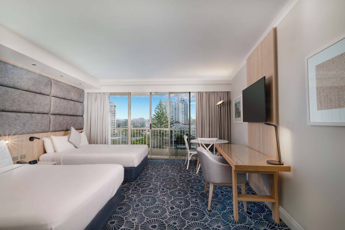 A hotel room with two beds, a desk, a chair, a TV, and a balcony with a city view.