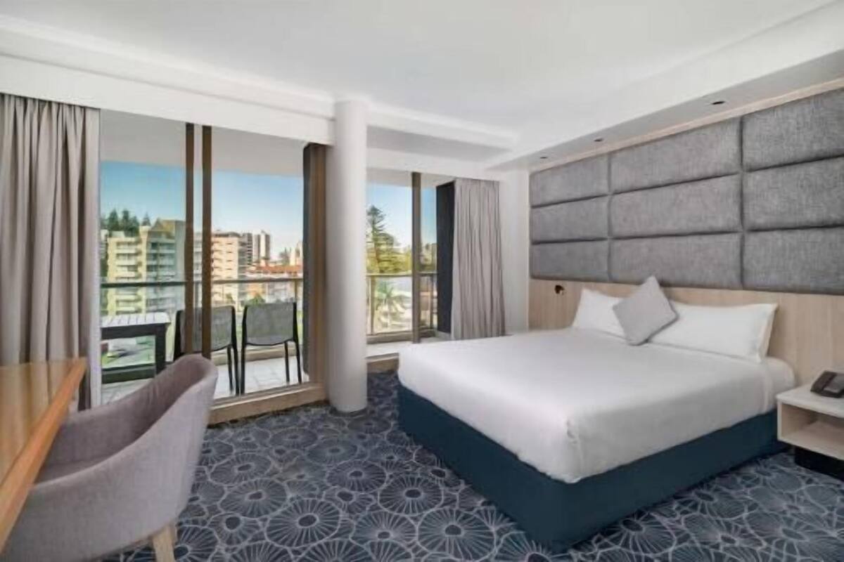 A hotel room with a bed, a desk, a chair, and large windows with a city view.