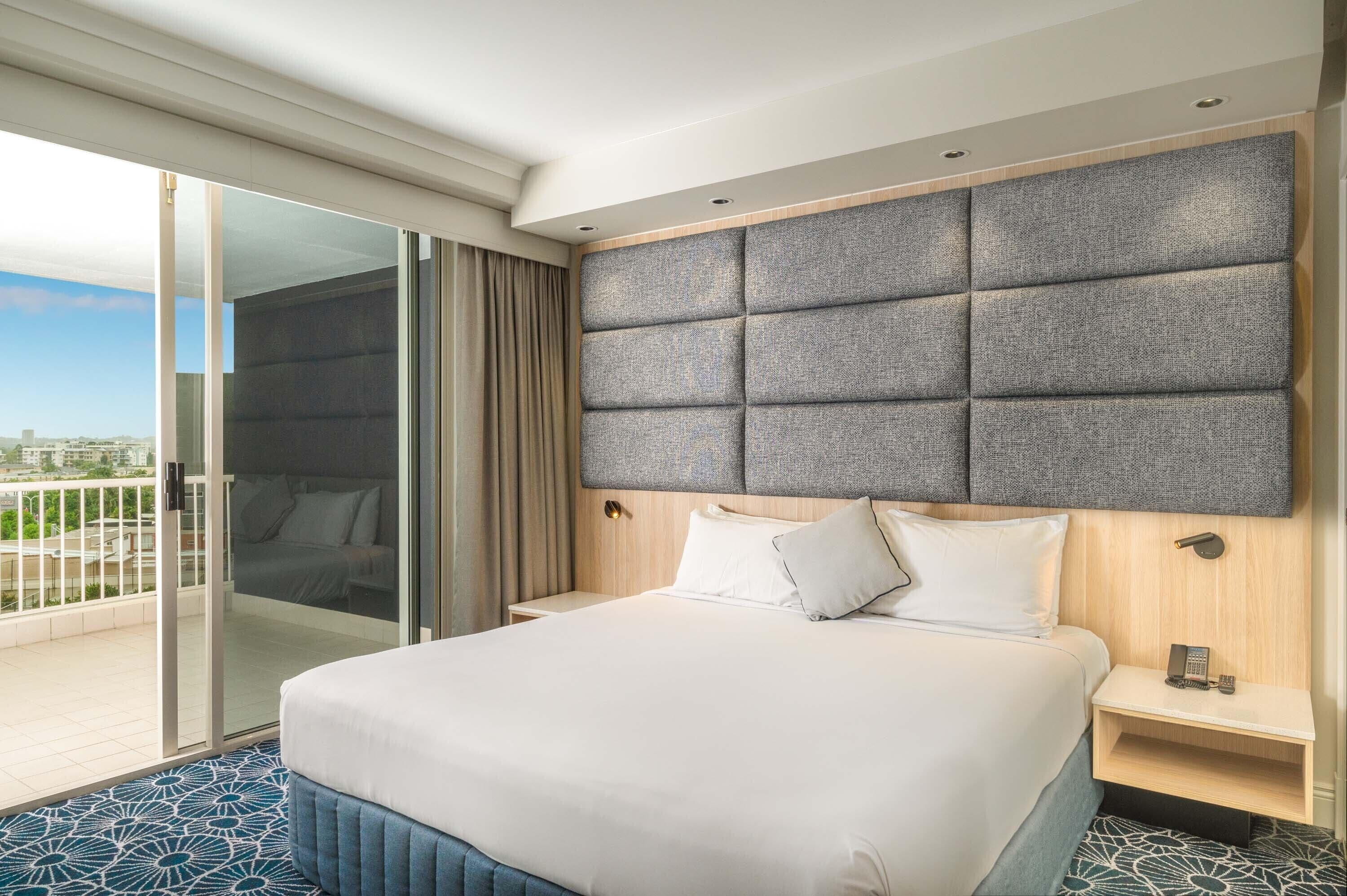 A hotel room with a large bed, a wooden headboard, a bedside table with a phone, and a sliding glass door leading to a balcony.