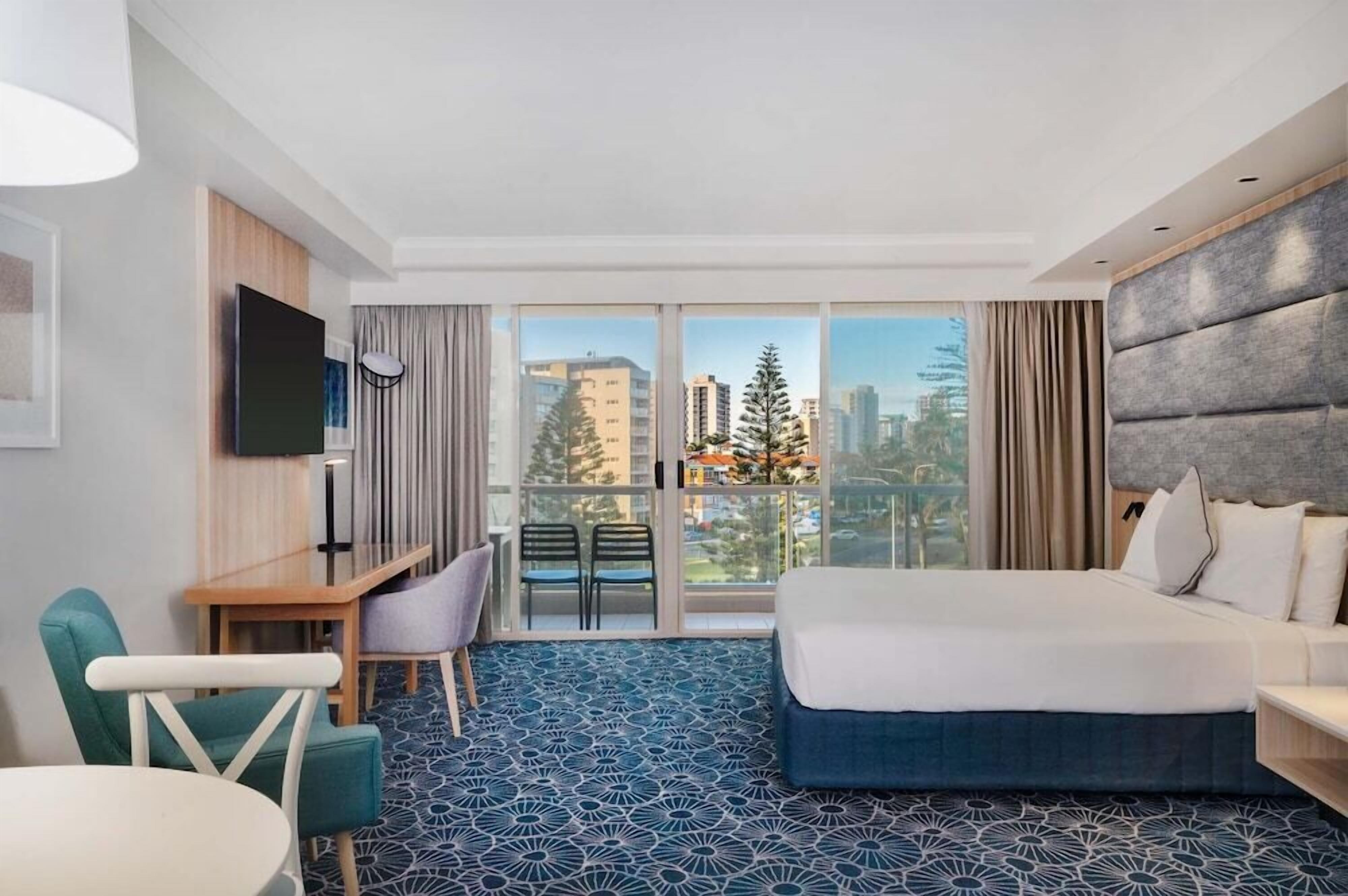 Ett hotellrum med en säng, ett skrivbord, en stol, en TV och ett stort fönster med utsikt över staden.