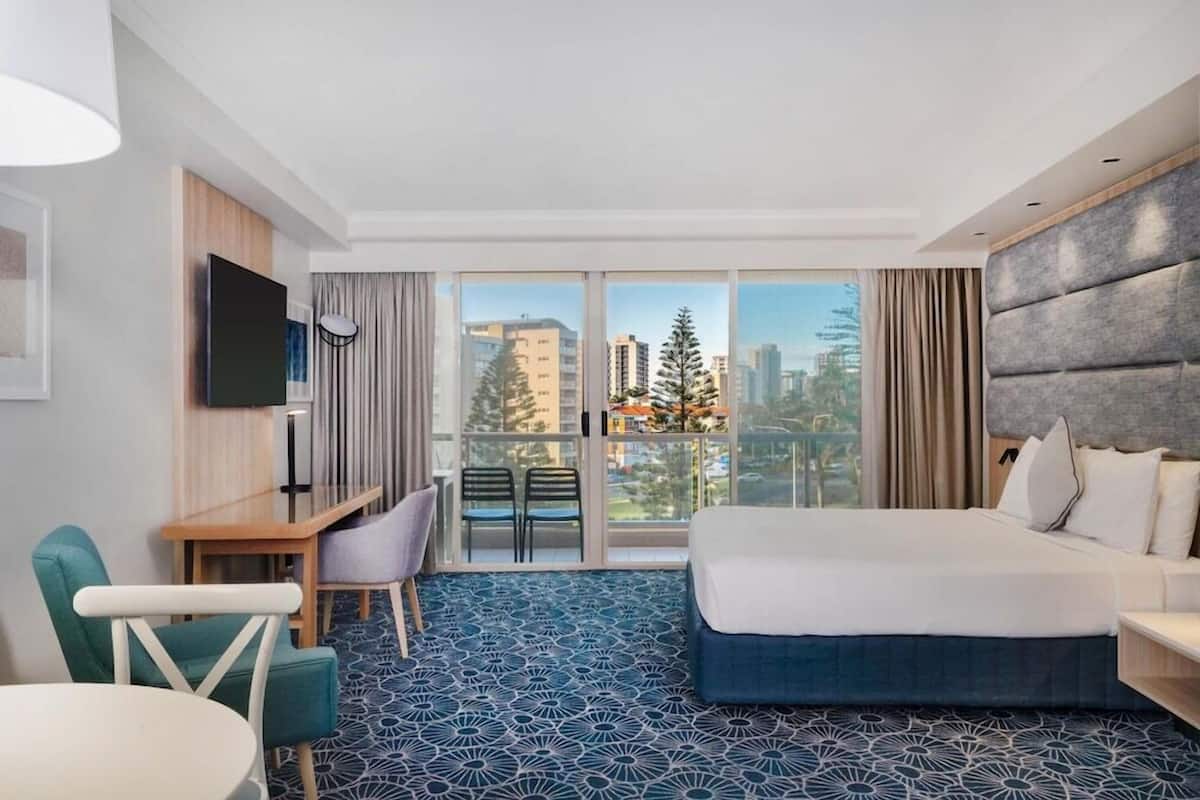 Ett hotellrum med en säng, ett skrivbord, en stol, en TV och ett stort fönster med utsikt över staden.