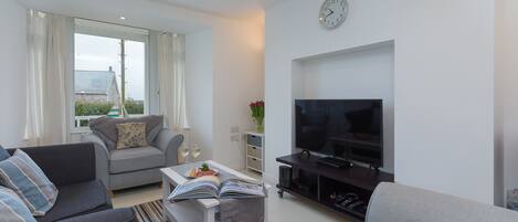 A modern living room with a sofa, a coffee table with a book, a flat-screen TV, and a window with curtains.
