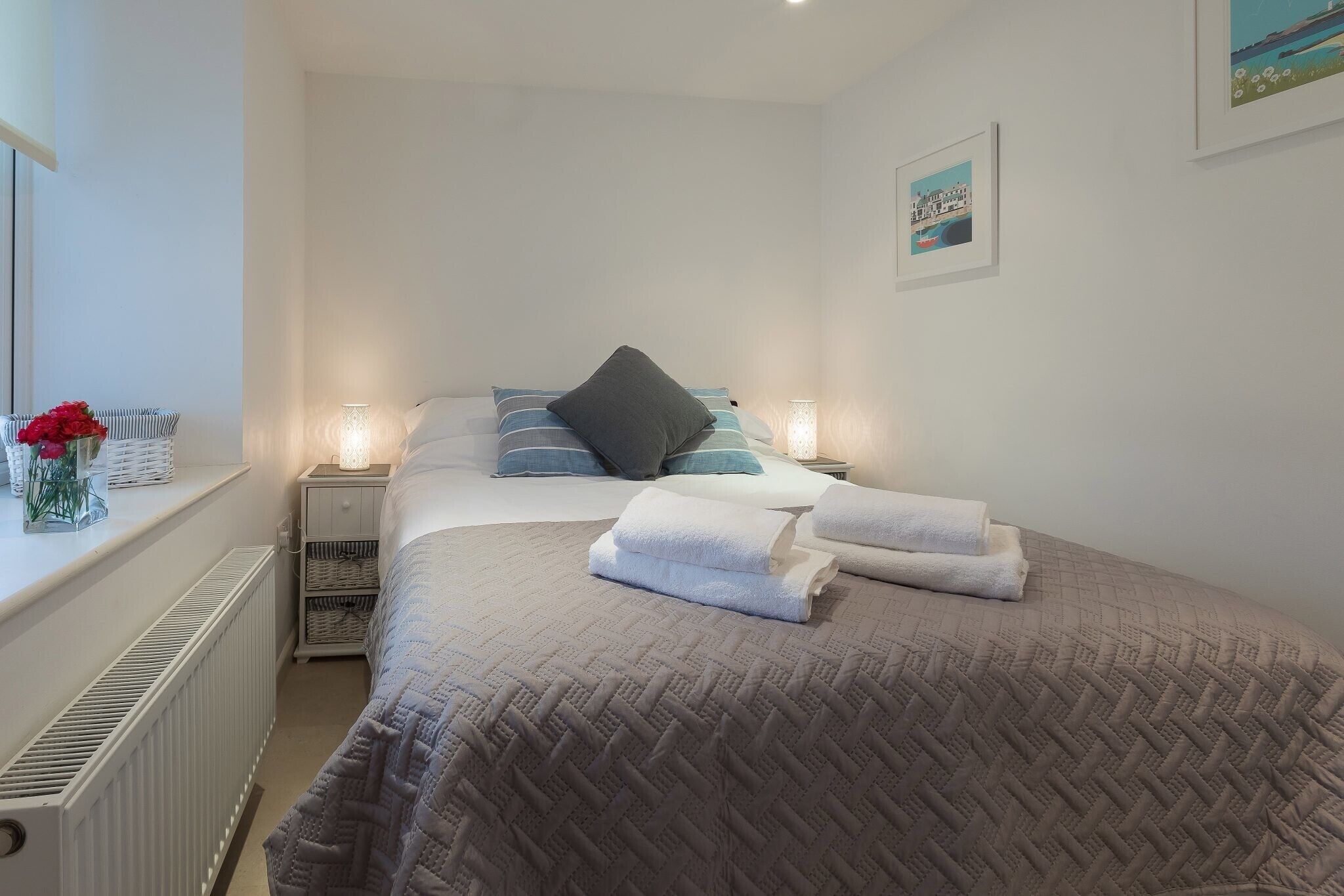 A neatly made bed with pillows and folded towels, a bedside table with a lamp, and a radiator on the wall.