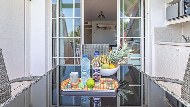 Een balkon met een glazen tafel, rieten stoelen, een fruitschaal, flessen en een dienblad.