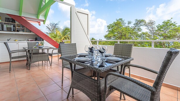 A terrace with wicker furniture and a dining table set for four, a kitchen area with a red staircase, and a view of trees and sky. - Studio Karuker’As - Aouara 1326 (Sainte-Anne)