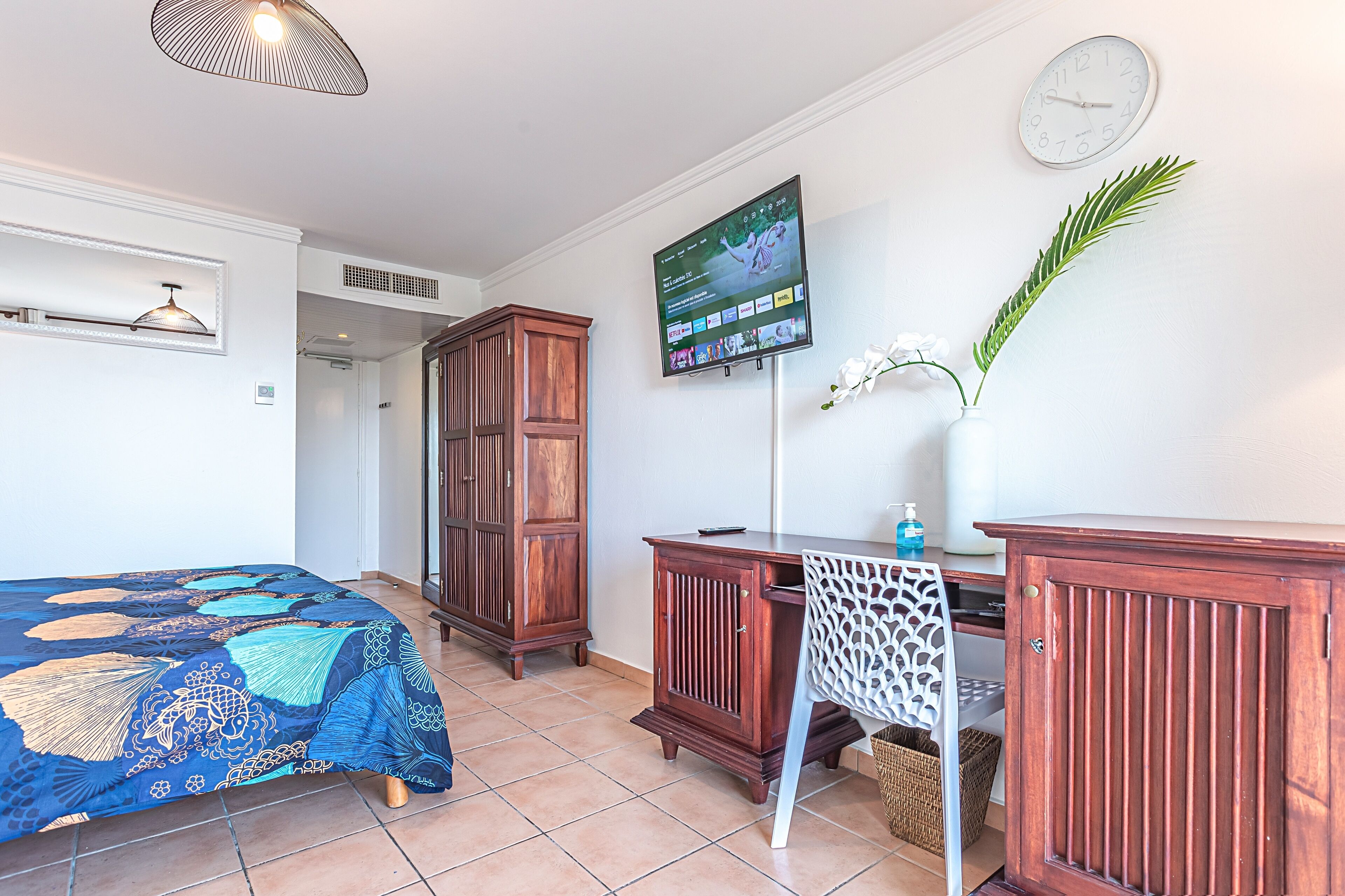 A room with a bed, a desk, a television, a clock, and a plant.