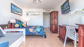 A bedroom with a bed, bedside table, lamp, and a TV on the wall. - Studio Karuker’As - Aouara 1326 (Sainte-Anne)