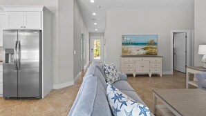 Una cocina moderna con un refrigerador de acero inoxidable, un comedor con mesa y sillas, un sofá gris con cojines estampados con estrellas, y un cuadro de una escena de playa en la pared.