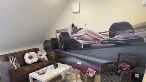A living room with a large racing car mural on the wall, a brown sofa, a white table, a wicker chair, and a small basket of books. - Beautiful vacation apartment at the pulse of the Green Hell (Kelberg)
