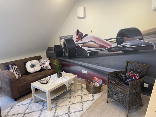 Una sala de estar con un gran mural de un coche de carreras en la pared, un sofá marrón, una mesa blanca, una silla de mimbre y una pequeña cesta con libros.